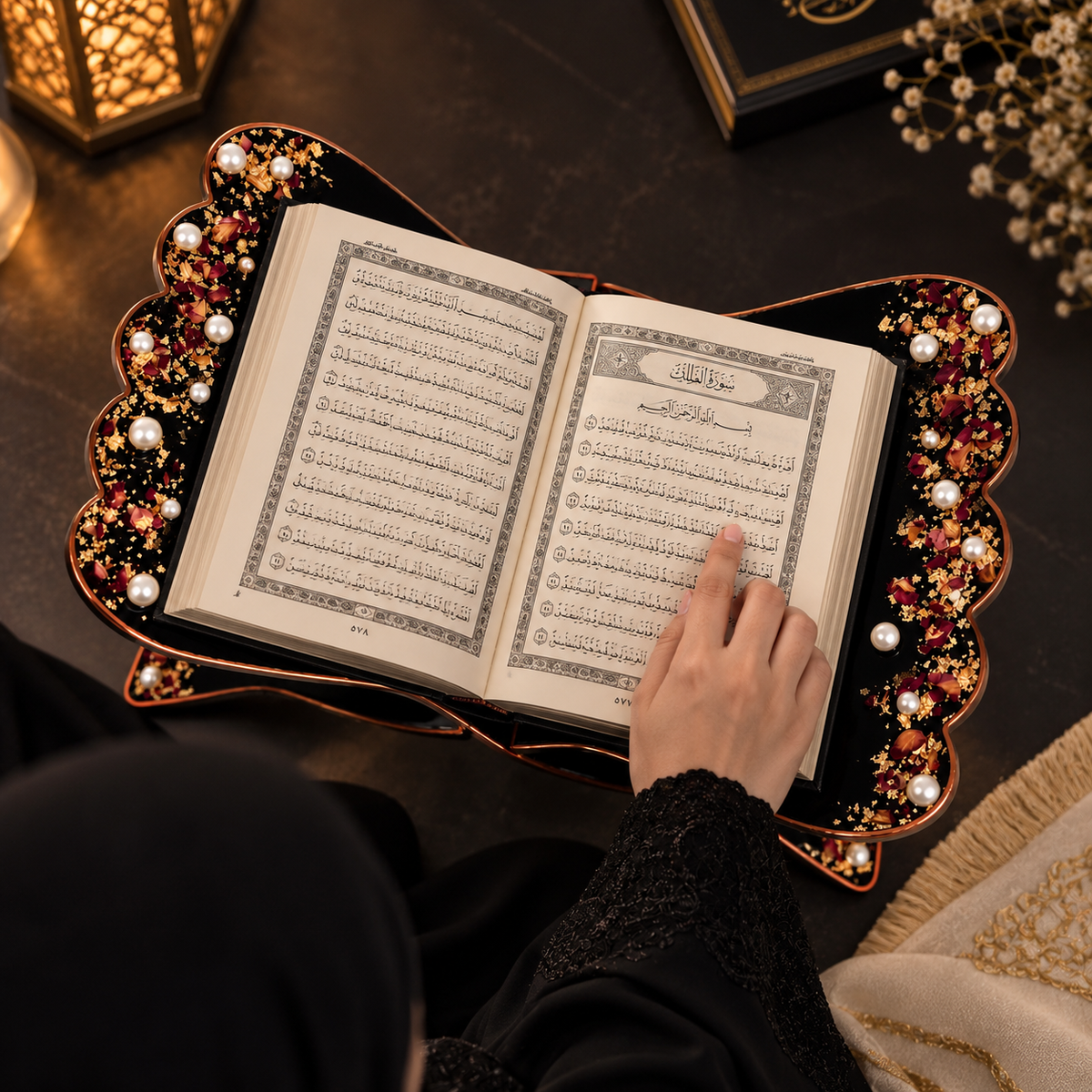 A premium black wooden Quran rehal with intricate gold detailing, pearl embellishments, and dried floral accents, placed on a dark elegant surface with warm ambient lighting and black satin fabric.
