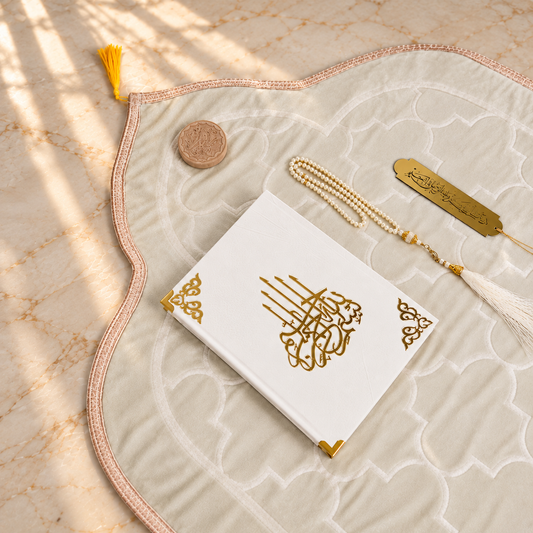 A styled flat-lay of a white Quran with gold Arabic calligraphy on the front cover, placed on a large cream-colored prayer mat with a pink border. The set includes a gold tassel bookmark, a gold Quran identification tag, a small wooden mezuzah case, and a white tassel, all arranged on a beige marble surface with soft window shadow overlays.