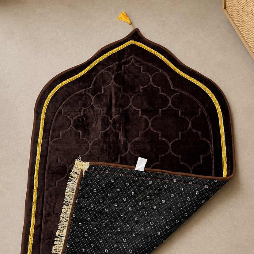 A dark brown and black prayer rug with gold trim and tassels, laid out on a beige tiled floor next to a wooden cabinet and a potted plant.