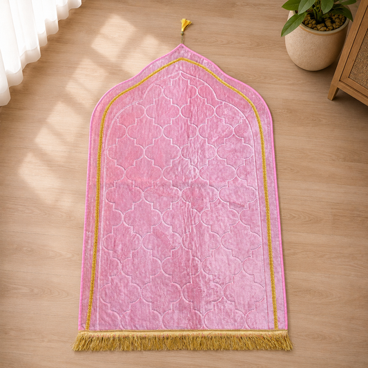 A pink prayer mat with a gold border and fringe, laid flat on a light wood floor next to a potted plant.