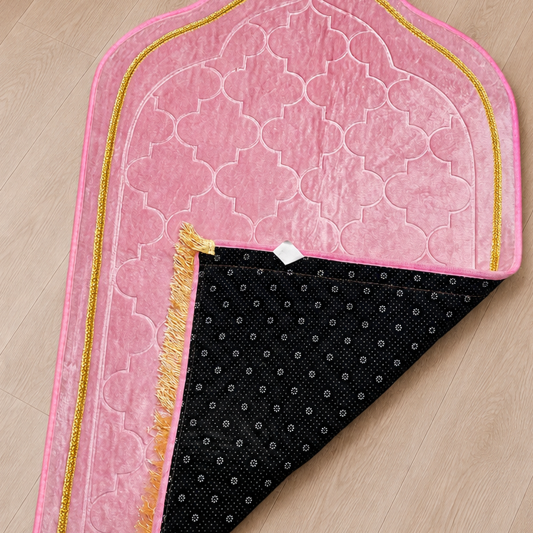 A pink prayer rug with gold trim and a black patterned underside, laid out on a light wood floor in a room corner with a vase and curtain visible.