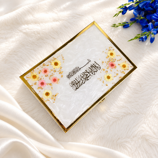 A rectangular white cosmetic compact with a gold border, decorated with small yellow and pink flowers and Arabic calligraphy, resting on a white faux fur surface with blue flowers in the corner.