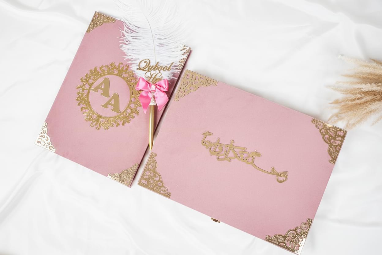 Pink invitation cards with gold text and a feather pen on a white surface