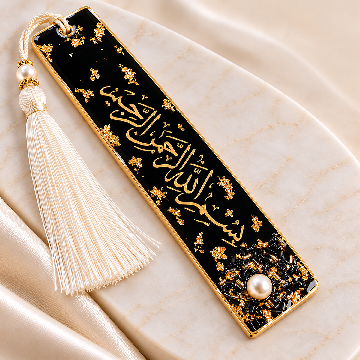 A rectangular black book bookmark with gold Arabic calligraphy, gold floral details, a cream tassel, and a pearl accent, displayed on a cream marble surface.