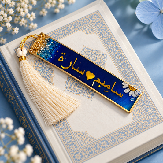 A rectangular enamel bookmark with a cream tassel lies on an open decorative book. The bookmark has a deep blue background with gold Arabic script, a small yellow heart, and a white flower with a gold center.