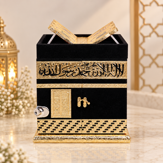 A decorative black and gold replica of the Kaaba prayer cabinet, shown on a marble surface with warm-toned decorative lanterns and flowers in the background.