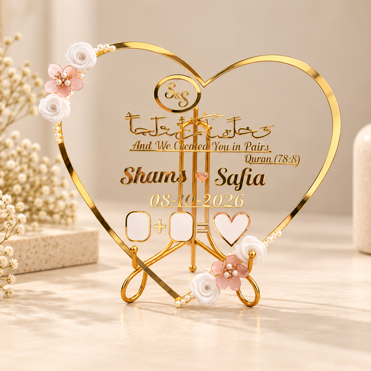 A gold heart-shaped acrylic and metal Islamic wedding plaque displayed on a light beige surface. The plaque features custom names 'Shams' and 'Safia', a date '08-10-2026', and placeholder boxes for dates and names, decorated with white and pink rose accents and pearl-like beads.