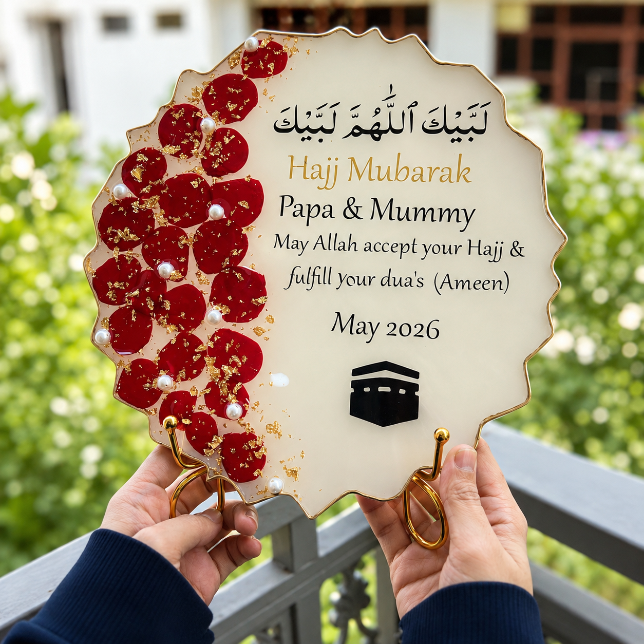 Hajj Mubarak stand with red floral design held by hands against a blurred green outdoor background