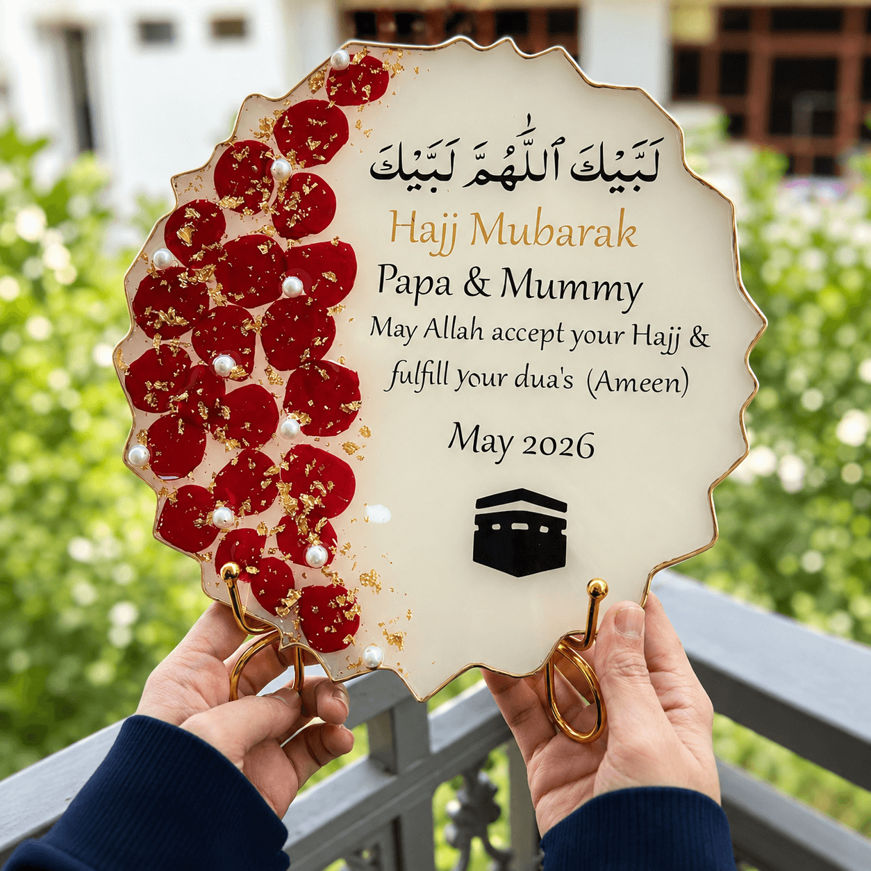 Hajj Mubarak stand with red floral design held by hands against a blurred green outdoor background