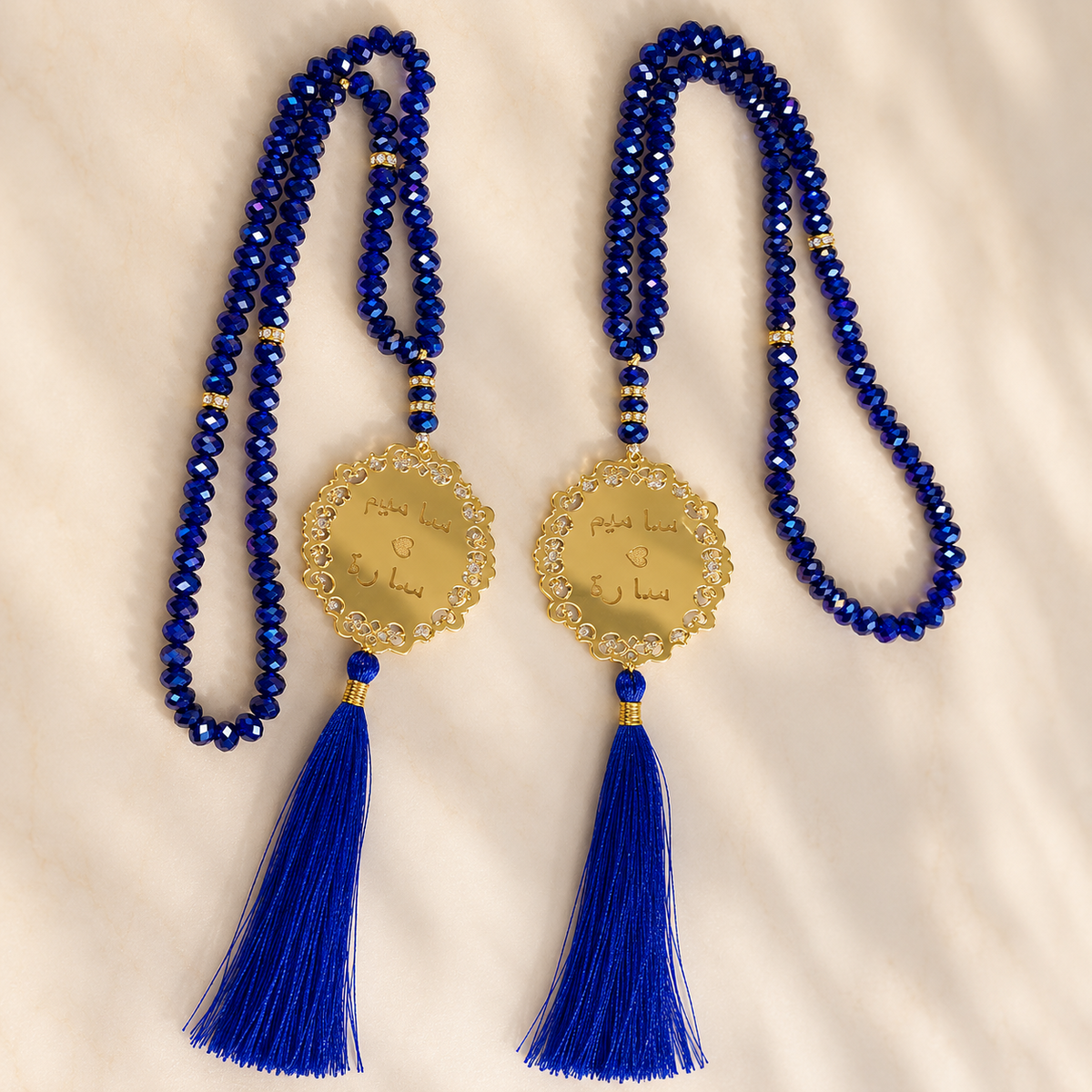 Two matching blue beaded prayer necklaces with gold medallions and blue tassels, laid flat on a light cream fabric background.