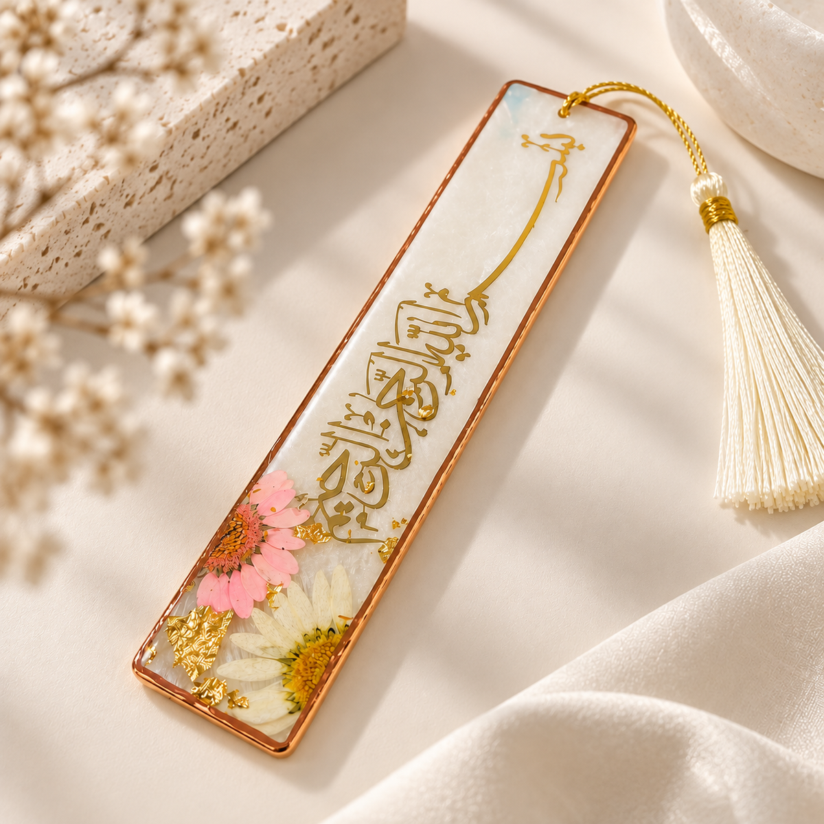 A rectangular resin bookmark with a copper-toned border, gold calligraphy, and pressed flowers, shown on a soft cream fabric with a tassel and dried flowers as styling props.