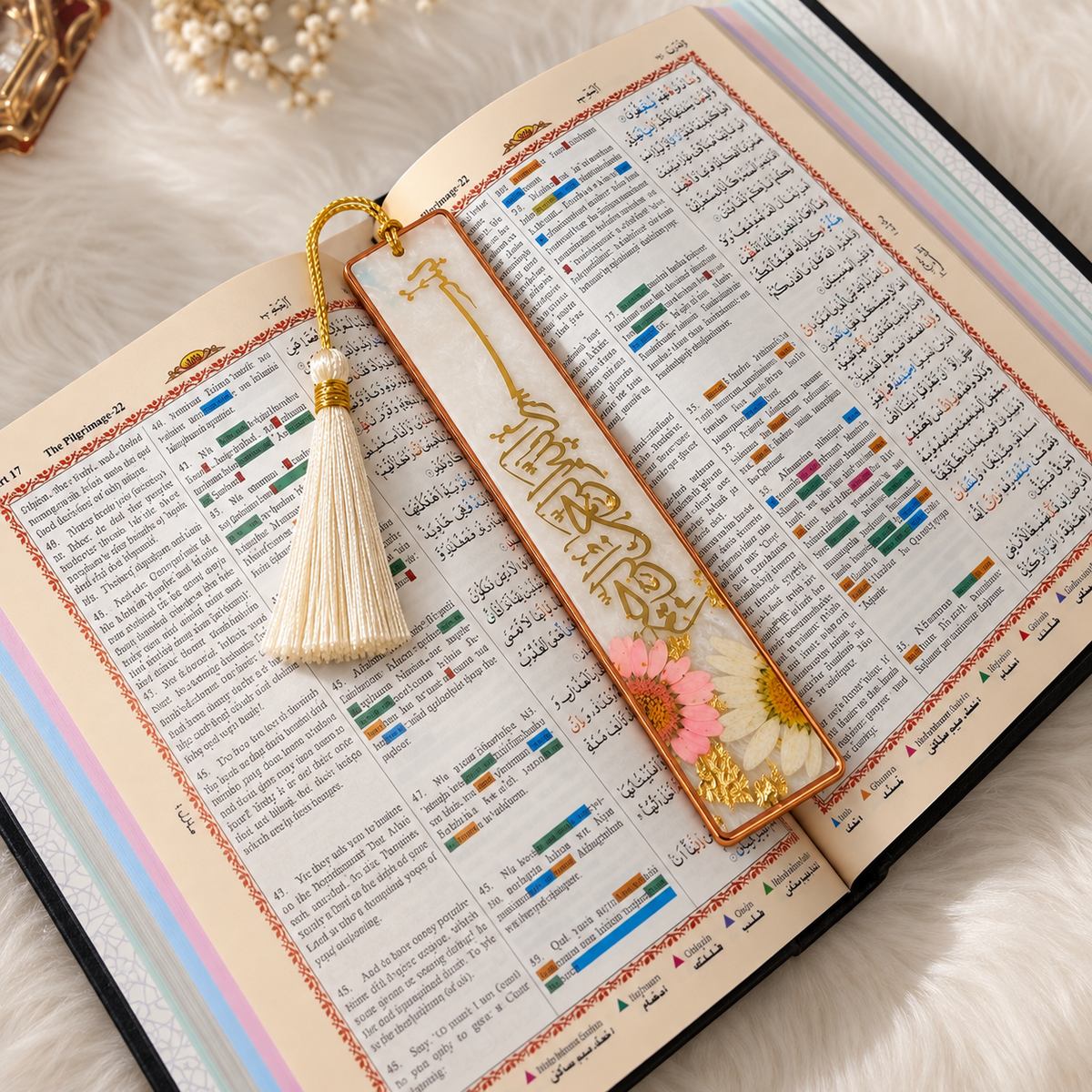 A decorative rectangular bookmark with gold Arabic calligraphy, a cream tassel, and dried pink and white flowers rests on an open book with printed text.