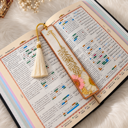 A decorative rectangular bookmark with gold Arabic calligraphy, a cream tassel, and dried pink and white flowers rests on an open book with printed text.