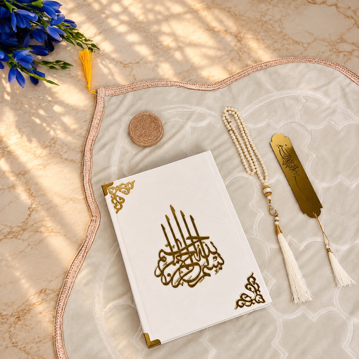 A white Quran with gold-embossed Arabic calligraphy and corner motifs, arranged on a beige patterned prayer mat with a tassel bookmark, a beaded tasbih, and a small coin. Blue flowers and warm sunlight shadows are visible in the background.