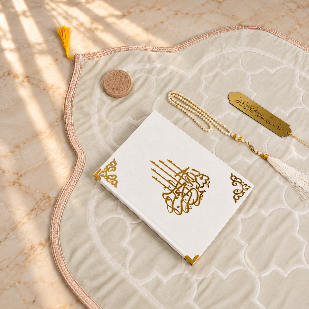 A styled flat-lay of a white Quran with gold Arabic calligraphy on the front cover, placed on a large cream-colored prayer mat with a pink border. The set includes a gold tassel bookmark, a gold Quran identification tag, a small wooden mezuzah case, and a white tassel, all arranged on a beige marble surface with soft window shadow overlays.