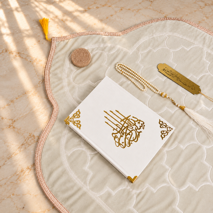 A styled flat-lay of a white Quran with gold Arabic calligraphy on the front cover, placed on a large cream-colored prayer mat with a pink border. The set includes a gold tassel bookmark, a gold Quran identification tag, a small wooden mezuzah case, and a white tassel, all arranged on a beige marble surface with soft window shadow overlays.