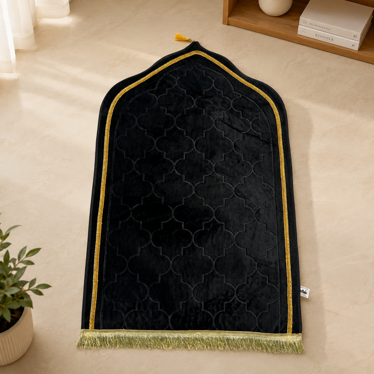 A black velvet prayer rug with a gold geometric trellis pattern, gold trim, and a gold fringe, laid out on a beige floor.