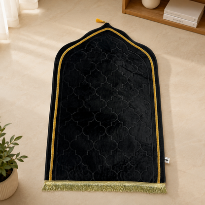 A black velvet prayer rug with a gold geometric trellis pattern, gold trim, and a gold fringe, laid out on a beige floor.