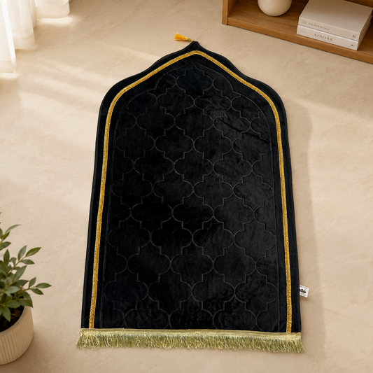 A black velvet prayer rug with a gold geometric trellis pattern, gold trim, and a gold fringe, laid out on a beige floor.