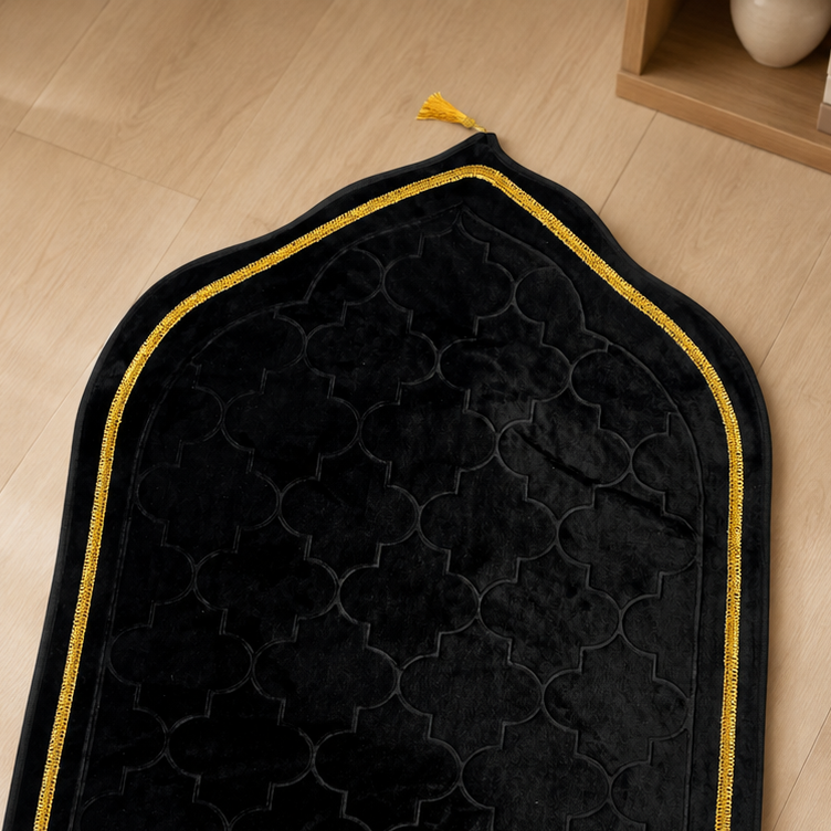 A black prayer rug with a subtle geometric pattern, gold trim, and a fringed bottom edge, laid out on a light wood floor.