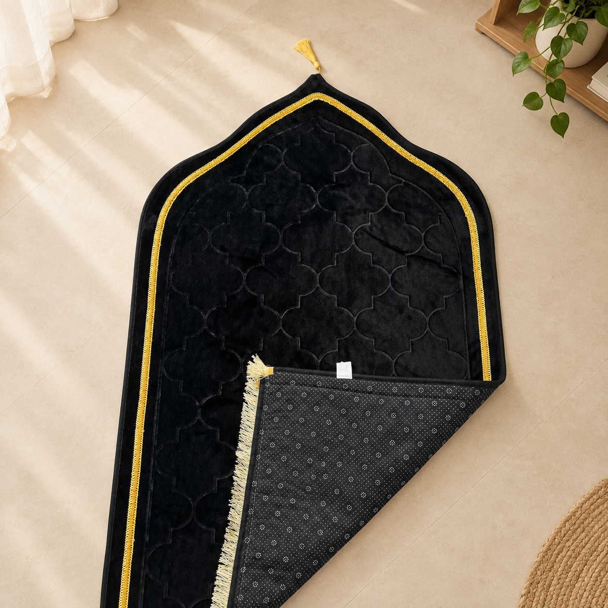 A black prayer rug with a gold embroidered border and a small tassel, laid flat on a beige floor with one corner folded over to show the patterned backing.