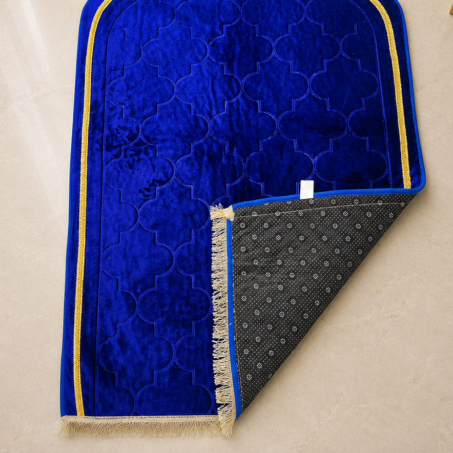 A royal blue velvet prayer mat with gold trim and a patterned gray underside, laid flat on a beige tiled floor with decorative props in the corners.