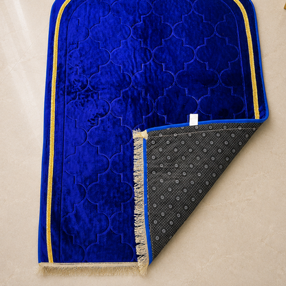 A royal blue velvet prayer mat with gold trim and a patterned gray underside, laid flat on a beige tiled floor with decorative props in the corners.
