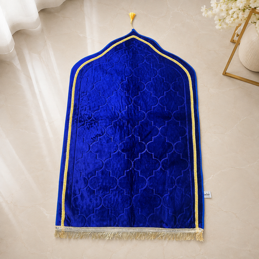 A royal blue velvet prayer rug with an arched shape, featuring a gold geometric border, a gold tassel at the top, and a gold-and-white fringed bottom edge, laid flat on a beige marble surface.
