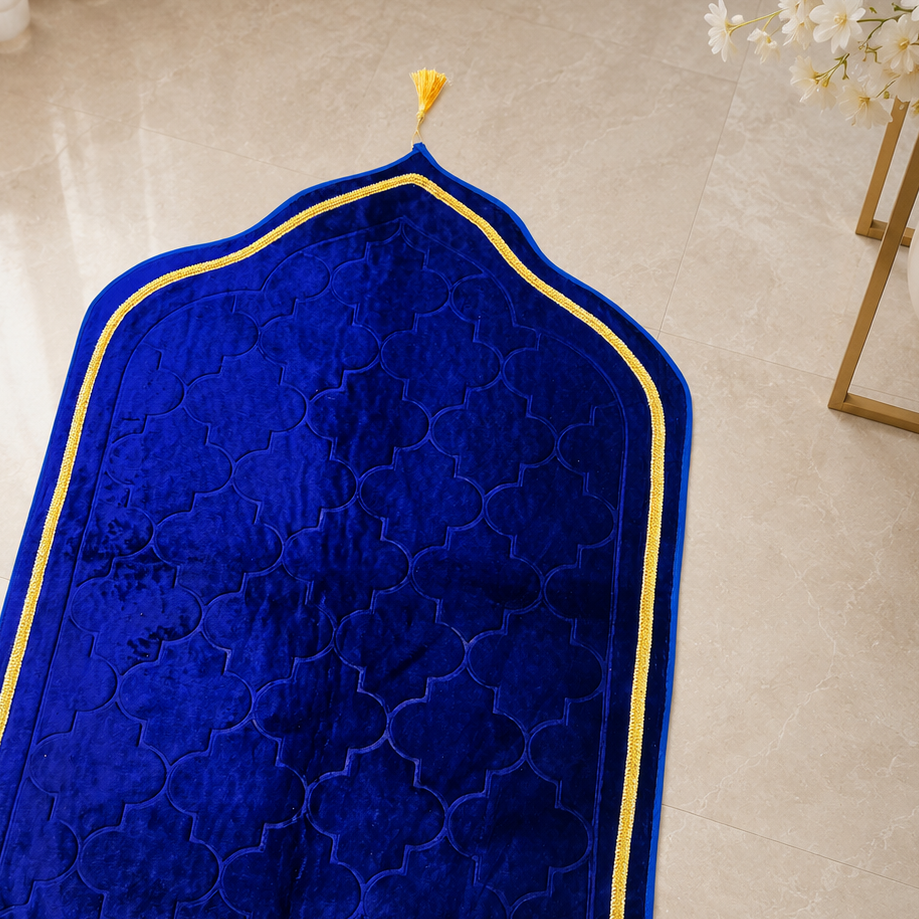 A royal blue velvet prayer rug with an arched top, gold embroidered border, and gold fringe, laid out on a beige marble floor.