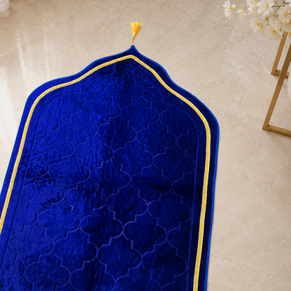 A royal blue velvet prayer rug with an arched top, gold embroidered border, and gold fringe, laid out on a beige marble floor.