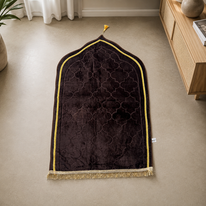 A dark brown velvet Islamic prayer mat with a gold geometric pattern, gold border trim, and a small gold tassel, laid out on a beige carpet in a modern room.