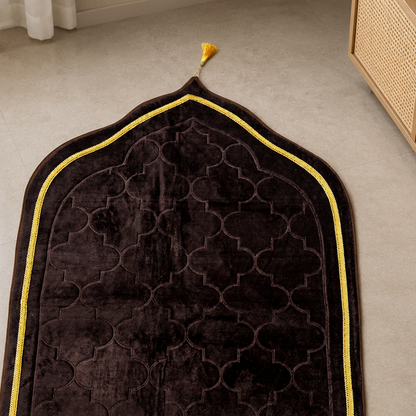 A dark brown velvet prayer rug with a geometric trellis pattern, a yellow border, and a matching yellow tassel and fringe, laid out on a beige tiled floor.