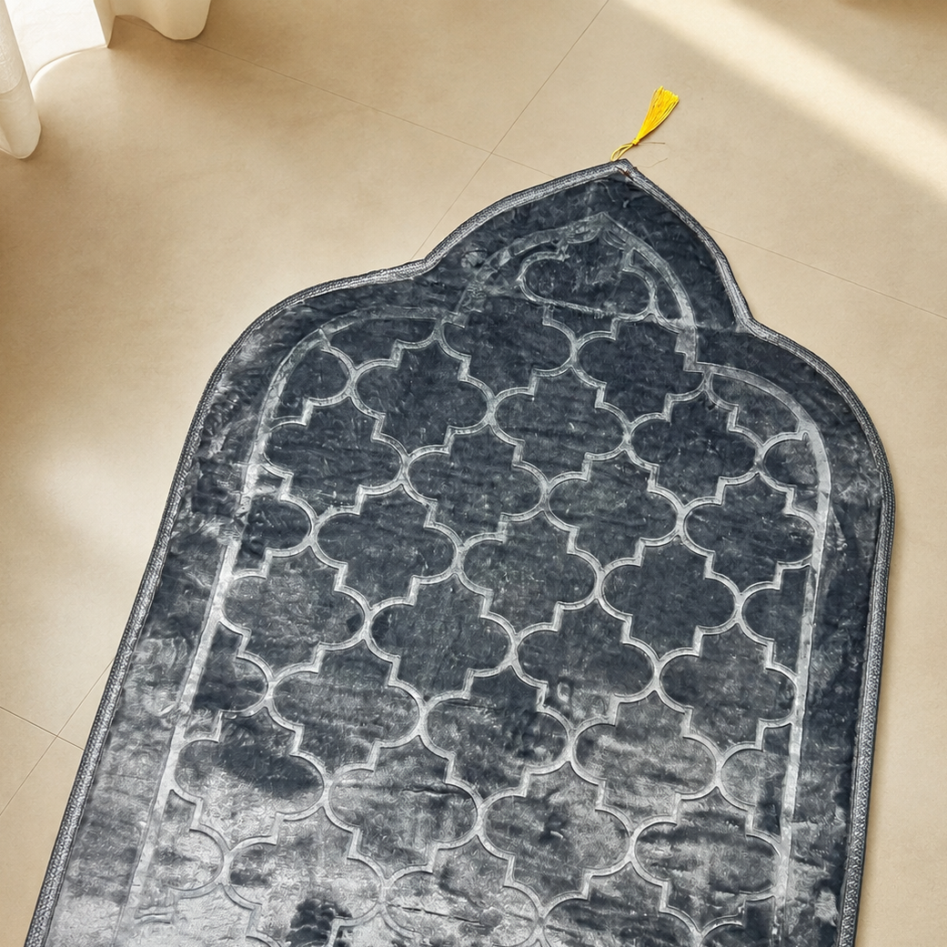 A dark gray and black prayer rug with a white Moroccan trellis pattern, laid out on a beige tiled floor.
