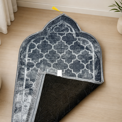 A dark blue and gray patterned prayer rug with a black reverse side, shown partially folded on a light wood floor in a styled room setting.