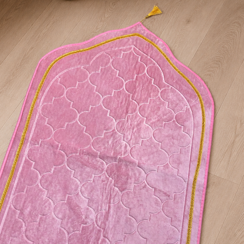 A pink prayer rug with a raised geometric pattern and a gold border, laid out on a light wood floor next to a white curtain and a small vase.
