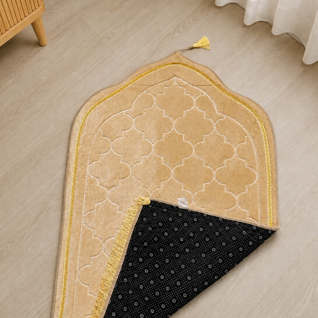 A beige and gold prayer rug with a geometric trellis pattern and tassels, laid out on a light wood floor. The underside of the rug is black with a small white geometric pattern.