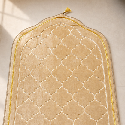 An arched beige prayer rug with a cream quatrefoil pattern, yellow-gold border, and matching fringed edge, laid on a light beige floor with soft window shadows.