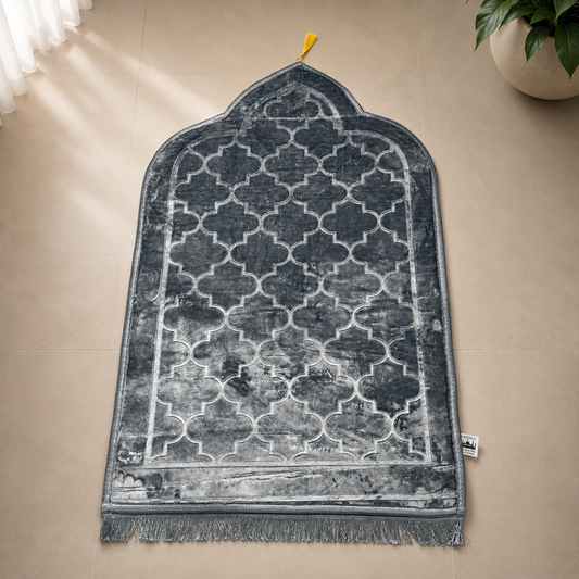 A dark gray and white Moroccan-style prayer rug with a trellis pattern and fringe, laid out on a beige tiled floor next to a potted plant.