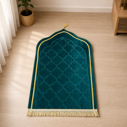 A teal green velvet Islamic prayer mat with a gold geometric border and cream fringe, laid out on a light wood floor in a sunlit room.