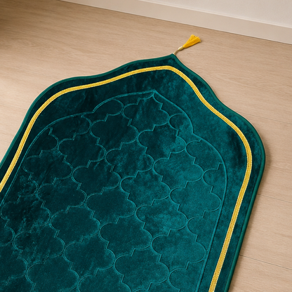 A dark teal green prayer rug with a geometric pattern, yellow border trim, and cream fringe, laid out on a light wood floor in a sunlit room.