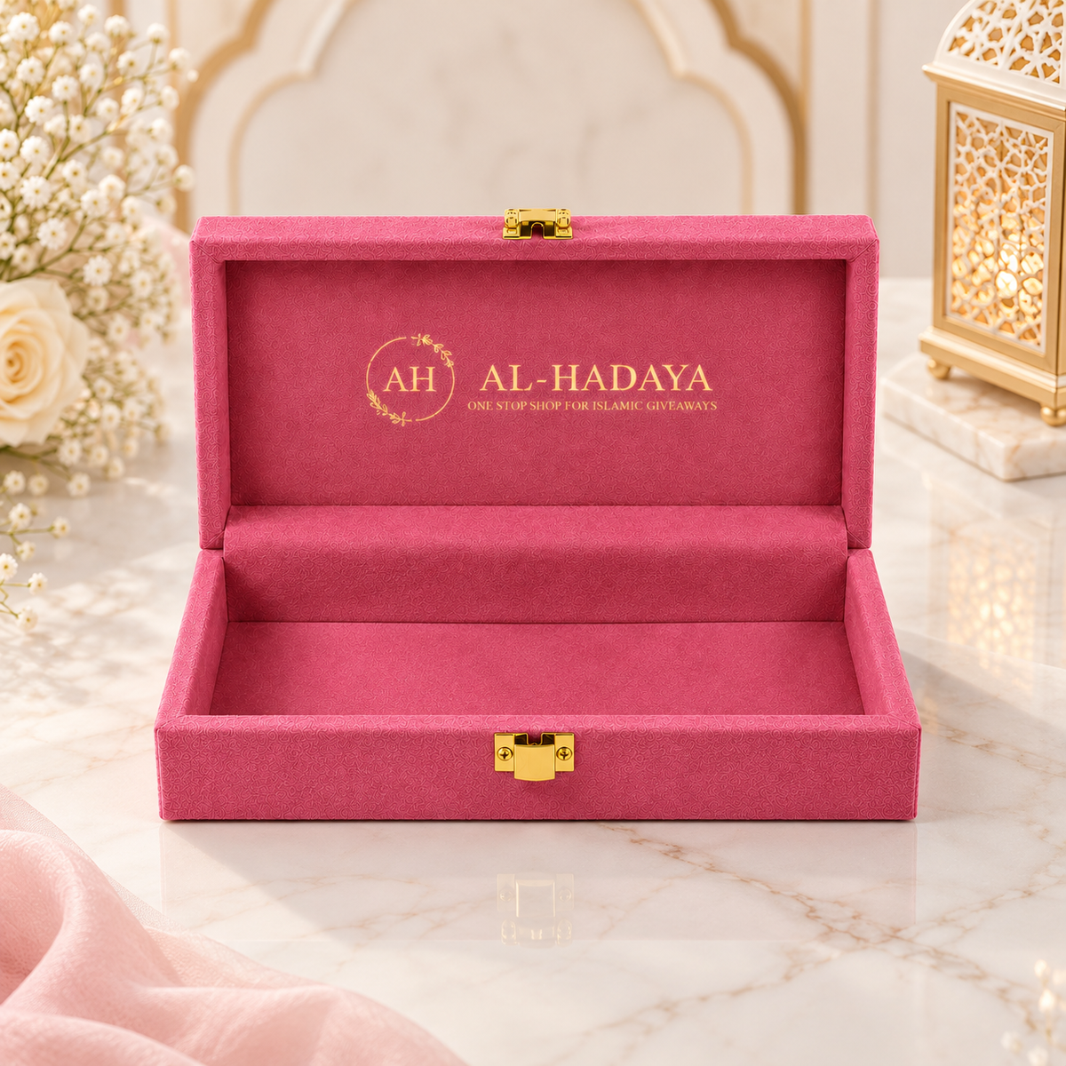 An open magenta pink velvet jewelry box with gold hardware, featuring the brand name 'AL-HADAYA' printed in gold on the inner lid, set on a marble surface with decorative flowers and a lantern in the background.