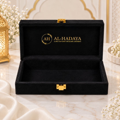 An open black velvet jewelry box with gold-tone hardware, featuring the brand name 'AL-HADAYA' printed in gold on the inside lid, displayed on a marble surface with decorative floral arrangements.