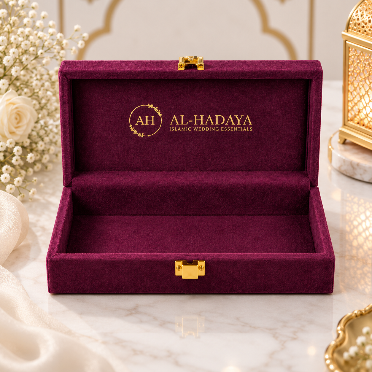 An open deep burgundy velvet jewelry box with gold-tone hardware, featuring the brand name 'AL-HADAYA' printed in gold on the inside lid, styled on a marble surface with decorative flowers and a lantern.