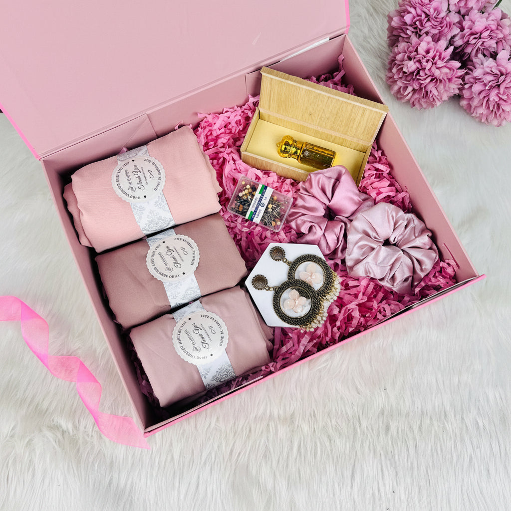 A pink gift box filled with pink hair accessories and a small wooden box containing a gold-tone hair pin, arranged on a white faux-fur surface with pink flowers in the corner.