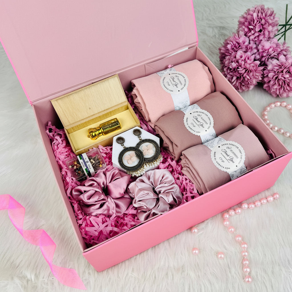 A pink gift box filled with a hair care set, including three rolled pink hair caps, a pair of black and gold hoop earrings, three pink satin scrunchies, a small wooden box with a gold bottle, and a small jar of hair pins, arranged on a white faux fur surface with pink flowers in the corner.