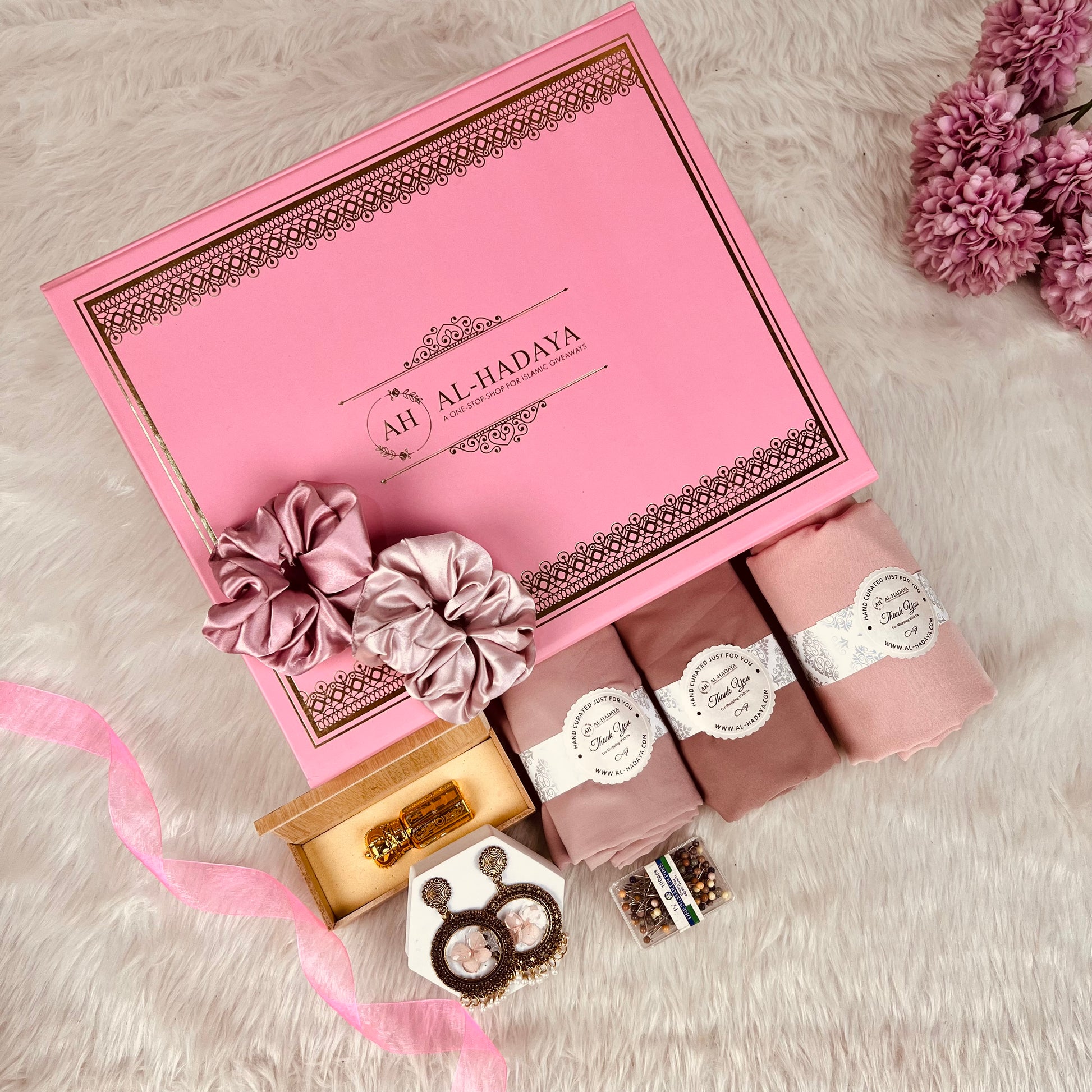A pink gift box set labeled 'AH AL-HADYA' arranged on a white faux-fur surface with pink flowers. The set includes three rolled pink items with white labels, a small gold box, a pair of round brown items, and pink ribbon.