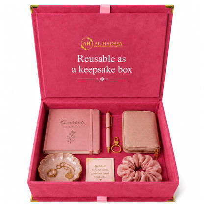 An open pink keepsake gift box with gold trim, branded 'AH AL-HADAYA', containing a pink journal, matching pen, zip wallet, small jewelry dish with gold-tone earrings, a card, and a pink scrunchie. The inside lid reads 'Reusable as a keepsake box'.