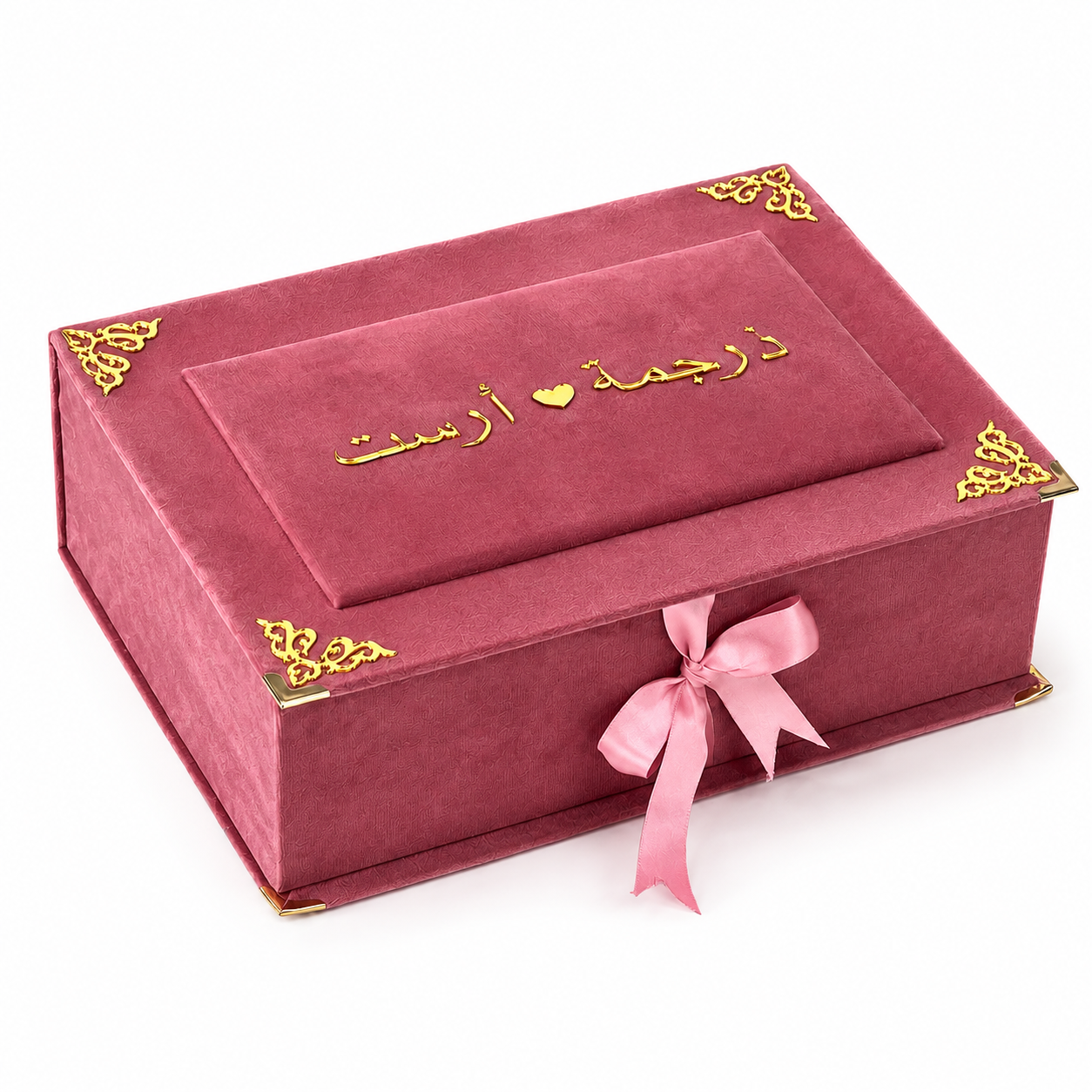 A rectangular pink velvet gift box with gold corner accents and gold Arabic calligraphy on the lid, tied with a pink ribbon bow, on a white background.