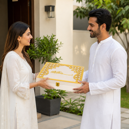 A man and a woman in white traditional clothing exchange a white gift box with ornate gold detailing and Arabic calligraphy, set in a modern outdoor patio environment.
