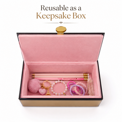 An open pink velvet keepsake box with a black border and gold clasp, shown on a white background with the text 'Reusable as a Keepsake Box' above it. Inside, pastel jewelry accessories including a pink pom-pom, beaded keychain, rings, bracelets, and a pencil are arranged.